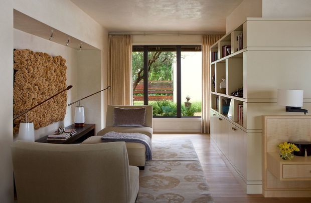Warm, refined living room with two upholstered lounge chairs, a sculptural textured wall panel, slender floor lamps, and a built-in cream-toned shelving and cabinetry unit. A large glazed opening with curtains looks onto a landscaped outdoor area, while a patterned area rug and timber-toned floor add softness and warmth.