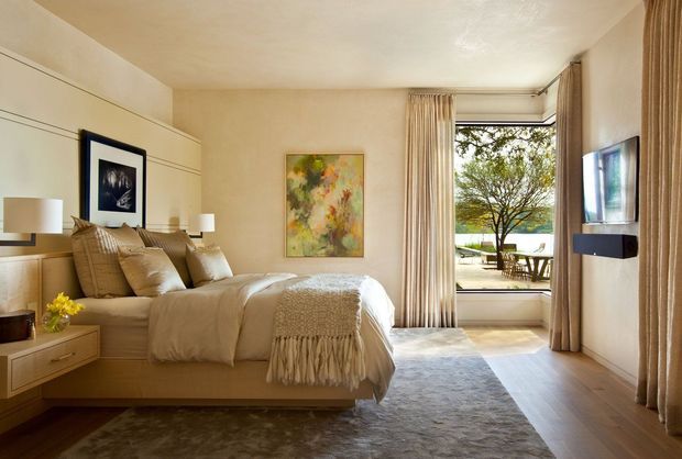 Warm, refined bedroom interior with a large upholstered bed, built-in bedside joinery, neutral bedding, floor-to-ceiling curtains, a wall-mounted television, framed artwork, and a large rug. Full-height glazing opens to an outdoor terrace or garden with visible greenery and seating. The room uses soft beige and cream tones with timber flooring and gentle natural light.