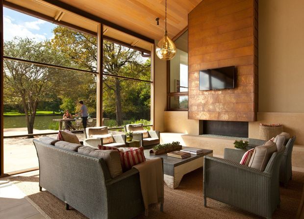 A warm, refined living room with floor-to-ceiling glazing overlooking a leafy garden and water feature. The room has a timber-lined ceiling, a large built-in fireplace clad in warm timber panels, a wall-mounted television, woven lounge chairs, a low rectangular coffee table, and neutral upholstered seating arranged around the fireplace.