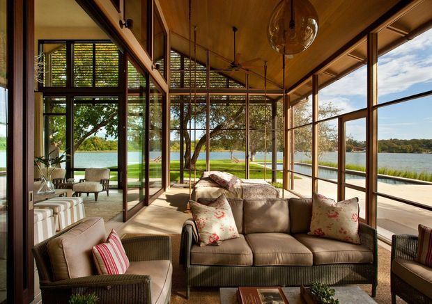 A sunlit enclosed living room or sunroom with extensive floor-to-ceiling glazing overlooking a calm waterfront landscape. The room features a warm timber-lined ceiling, dark timber-framed windows and doors, woven lounge furniture, a central sofa with floral cushions, and ceiling-hung pendant and fan fixtures. Reflections reveal additional seating and the room's glazed enclosure.