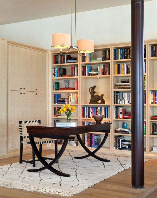 A refined study or library with floor-to-ceiling light timber built-in bookshelves and concealed cabinetry, filled with books and displaying a small sculpture. A dark wood trestle-style desk with curved black supports sits on a pale patterned rug, accompanied by a woven chair. A sculptural pendant light hangs overhead, and a dark cylindrical structural column is prominent in the foreground. The room has warm timber flooring and a calm, curated character.