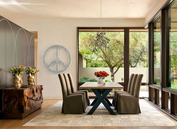 A refined dining area with a rectangular table and upholstered dining chairs, positioned beside large floor-to-ceiling windows overlooking a landscaped courtyard. The room features a sculptural dark wood sideboard, dark built-in wall panelling with curved line detailing, a textured peace-symbol wall feature, a light tiled floor, and warm neutral finishes. The extensive glazing creates a bright connection to the outdoors.