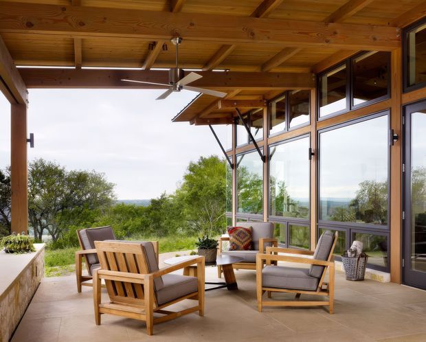 A covered outdoor living terrace with timber posts, exposed timber ceiling beams, a ceiling fan, and extensive glazed doors and windows. The space is furnished with a timber outdoor lounge setting, cushioned armchairs, a low table, and potted plants, overlooking a leafy landscape and distant horizon. The architecture has a warm, rustic timber character with large operable glazing and a strong indoor-outdoor connection.
