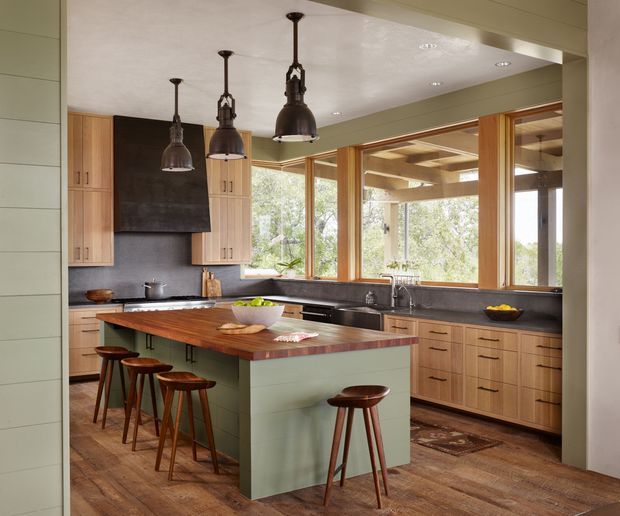 Warm, spacious kitchen with a large central timber island clad in muted sage-green cabinetry, integrated seating, and several timber stools. Light timber cabinetry lines the perimeter, with a dark stone or charcoal splashback and benchtop accents. Large timber-framed windows overlook trees and connect visually to a covered outdoor structure. Three oversized dark metal pendant lights hang over the island, while the timber flooring and natural materials create a refined rustic atmosphere.