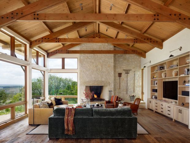 A spacious rustic living room with a dramatic exposed timber post-and-beam vaulted ceiling, warm timber flooring, and large windows overlooking a green landscape. The room includes a dark sectional sofa, lounge chairs, a stone fireplace with a lit fire, and extensive built-in shelving and media cabinetry.