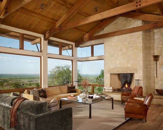 Spacious rustic living room with a vaulted timber ceiling and exposed post-and-beam structure. Large corner windows overlook a broad natural landscape, while a substantial light stone fireplace forms the main feature wall. The room contains sofas, leather armchairs, a glass coffee table, and a large neutral rug, creating a warm lodge-like retreat.