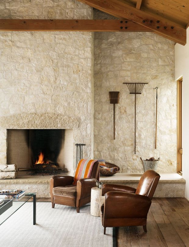 Rustic living room with a large exposed pale stone fireplace wall, built-in hearth seating, exposed timber roof beams and a pitched timber ceiling. Two brown leather lounge chairs face the fireplace, with a glass coffee table and neutral rug on a dark timber floor. Decorative objects and fire tools add a warm, lodge-like character.