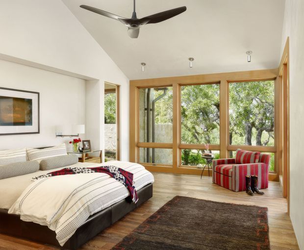 Bright contemporary bedroom with two low-profile beds dressed in white striped bedding, a red striped armchair, timber flooring, and large timber-framed windows overlooking greenery. A ceiling fan, framed artwork, bedside furniture, and a dark area rug complete the relaxed interior.