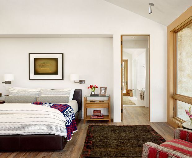 Bright, refined bedroom with a large upholstered bed dressed in patterned white bedding and a colourful throw, dark timber flooring, a bedside table, framed artwork, and wall-mounted reading lights. A timber-framed glazed window, patterned roller blind, and red woven armchair add warmth. An open doorway leads to a hallway or bathroom area.