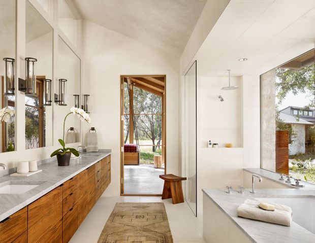 A bright, refined bathroom with a long timber vanity and pale stone benchtop, large wall mirror, twin wall-mounted sconces, freestanding basin fixtures, and a spacious white tiled wet area. Full-height glazing and an open doorway connect the room visually to a leafy outdoor setting. A small timber stool and runner add warmth.