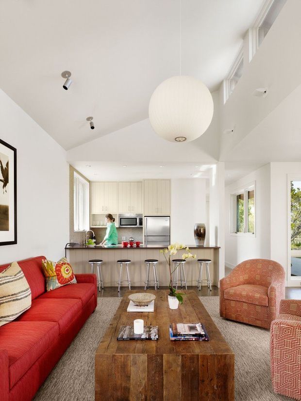 Bright open-plan living room with a vivid red sectional sofa, patterned upholstered armchairs, a large rustic timber coffee table, and a compact kitchen with island seating in the background. The room has white walls and ceilings, high-level glazing, recessed spotlights, and a large spherical pendant light, creating an airy, contemporary interior.