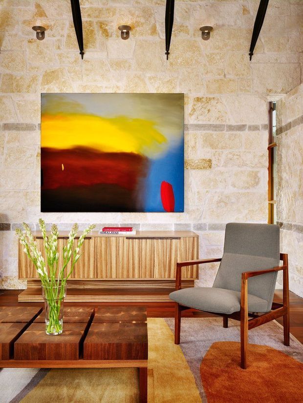 Warmly styled living room with exposed pale stone block walls, dark timber ceiling beams, a large abstract painting, mid-century timber credenza and coffee table, and a grey upholstered timber-framed armchair. A patterned rug, sculptural wall sconces, and a vase of greenery complete the space.