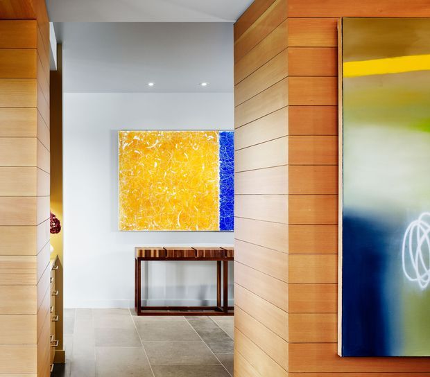 A bright contemporary interior hallway or entry passage framed by warm horizontal timber wall panelling and joinery. Large-format pale stone or porcelain floor tiles lead toward a slim timber console table beneath a vivid abstract artwork. A tall colourful artwork is partially visible on the right wall, with recessed ceiling downlights and clean white walls creating a refined gallery-like atmosphere.