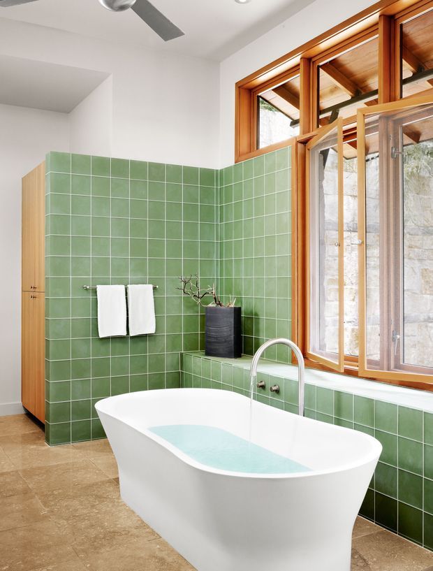A bright contemporary bathroom featuring a freestanding white bathtub, green square tiled walls and bath surround, a curved chrome tap, timber-framed opening windows, and warm timber ceiling and exterior structure visible beyond the glazing. The room has a tiled floor, built-in tiled ledge, and minimal white sanitary fittings.