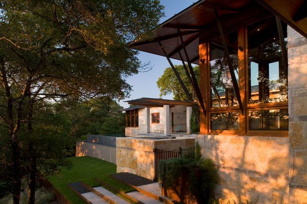 Exterior view of a contemporary multi-level residence set among mature trees on a sloped, landscaped site. The building features broad roof overhangs, extensive glazing, exposed dark structural framing, warm timber elements, and light natural-stone walls. Stepped stone paths and terraces connect the house to the garden, with a smaller flat-roofed wing visible behind.