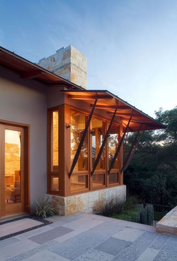 Exterior view of a contemporary house with a projecting glazed sunroom or enclosed terrace. The extension features warm timber-framed glazing, angled dark metal supports, and a broad sloping roof canopy. Pale stone veneer forms the base and a tall stone-clad masonry volume rises behind the lower roof. A paved terrace occupies the foreground, with landscaped vegetation beyond.