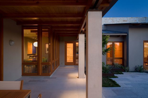 A covered residential entry courtyard or walkway at dusk, featuring warm timber-framed glazing and doors, exposed timber ceiling beams, pale rendered columns, and grey stone or concrete paving. The view looks toward a softly lit interior and an adjoining courtyard facade with warm amber windows, creating a sheltered, refined indoor-outdoor transition.