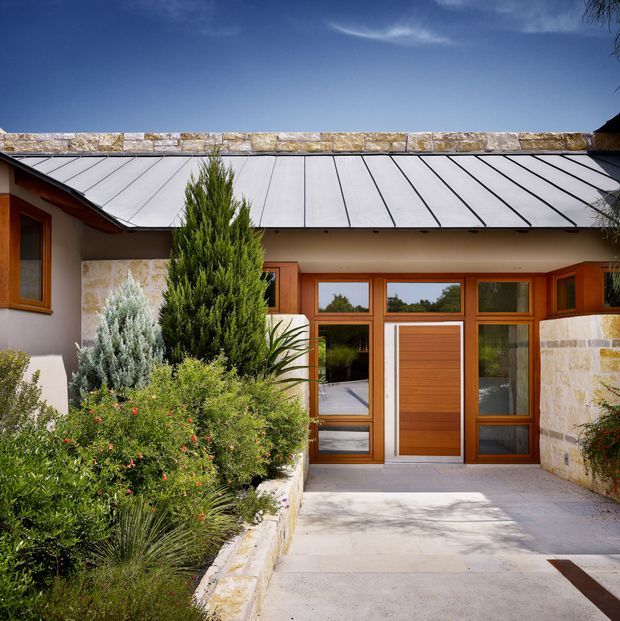 Front exterior view of a low-rise residential entry, featuring a standing-seam metal roof, pale stone masonry walls, rendered sections, and a timber-framed glazed entrance with a solid timber door. A landscaped garden bed and paved entry path lead toward the doorway under clear daylight.