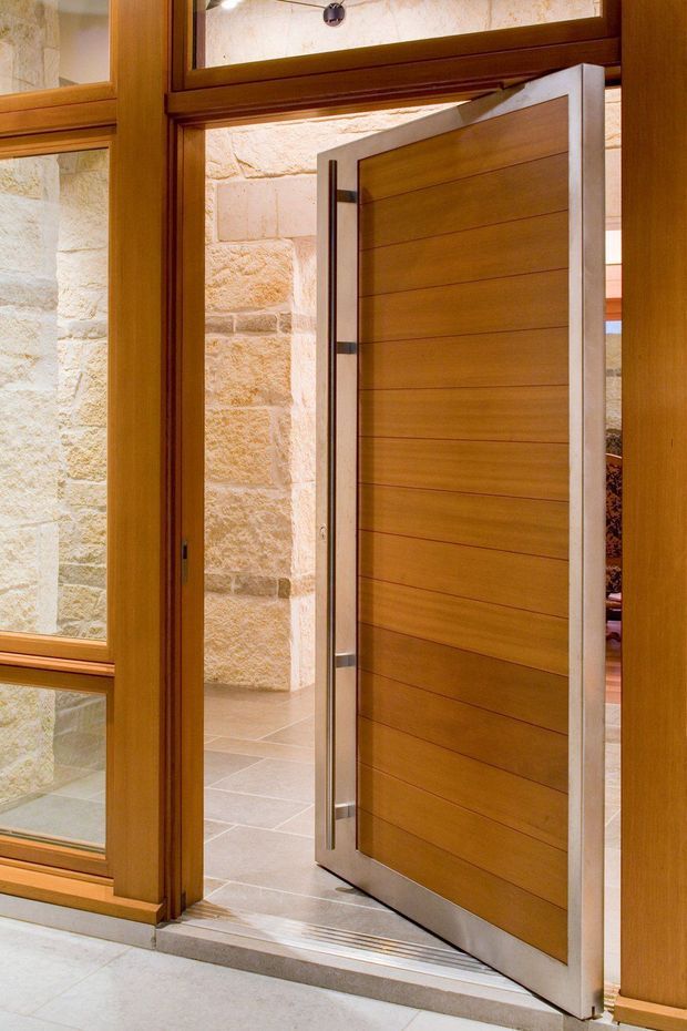 Close-up interior view of an open modern timber door with horizontal wood panelling and brushed metal edging. The doorway is framed by rich stained timber, with glazed timber sidelights and transom panels. A pale textured stone wall and light tiled floor are visible beyond, creating a warm, refined entry transition.