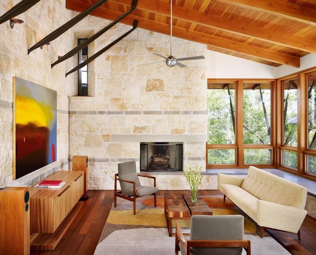 A warm, mid-century-inspired living room with a substantial natural stone fireplace wall, exposed timber pitched ceiling, dark structural beams, and extensive timber-framed glazing. The room contains a cream sofa, mid-century lounge chairs, a timber media console, a low timber coffee table, layered rugs, and a ceiling fan. Abundant windows overlook trees, creating a bright connection to the outdoors.