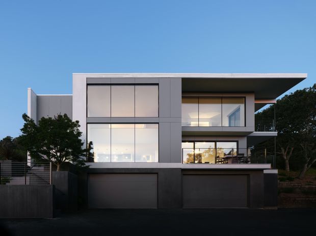 Front exterior view of a contemporary multi-storey residence at dusk, featuring a flat roof with broad cantilevered overhangs, dark grey rendered or panelled facade surfaces, expansive floor-to-ceiling glazing, and two integrated garage doors at ground level. Trees and boundary fencing frame the property, while warm interior lighting is visible through the windows.