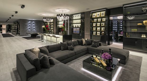 A spacious, sophisticated open-plan living and entertaining area with a large charcoal U-shaped sectional sofa, illuminated black glass coffee table, dark built-in display cabinetry, and a bar or kitchenette at the rear. The room features polished grey flooring, a light ceiling with recessed spotlights, dramatic chandeliers, dark wall finishes, and carefully arranged display objects and artwork. The overall scheme is high-contrast, dark, refined, and luxurious.