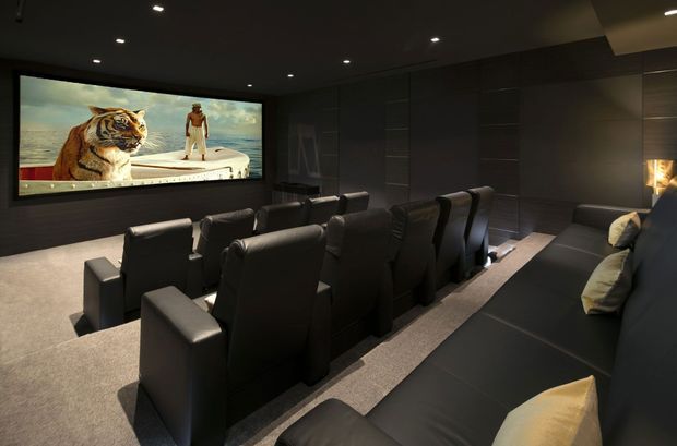 A dark, luxurious dedicated home cinema with multiple rows of black reclining theater seats, a large wall-mounted projection screen, dark paneled walls, recessed ceiling lighting, and a long black sofa along the side wall. The room has carpeted flooring and a moody, immersive atmosphere.
