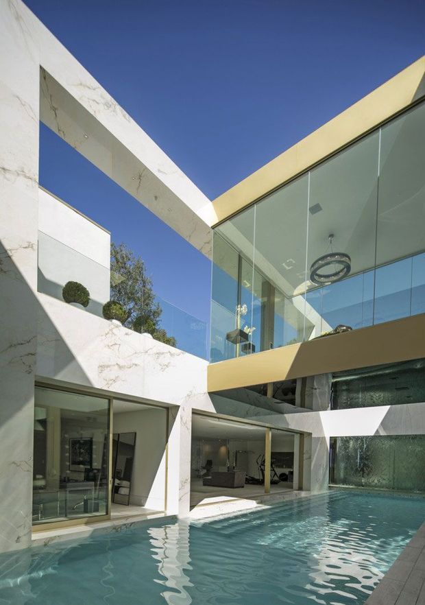 A dramatic contemporary multi-level courtyard interior/exterior space centred on a swimming pool. White marble-look walls and monumental rectangular openings frame the view, while expansive floor-to-ceiling glazing reveals an upper-level living area with a circular pendant light. Strong geometric forms, reflective glass, a gold-toned horizontal fascia, sculpted topiary, and crisp sunlight create a luxurious modern architectural composition.
