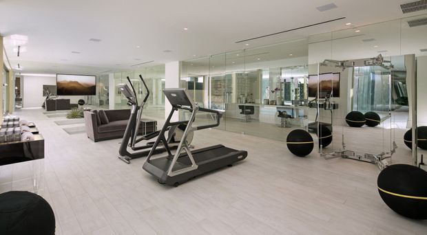 Bright, spacious contemporary home gym with mirrored walls, pale wood-look flooring, cardio equipment, black exercise balls with yellow bands, and a lounge area visible at the rear. Recessed ceiling lighting and extensive mirrors create an airy, polished fit-out.