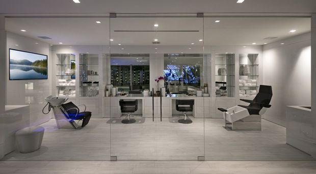 A sleek, spacious contemporary bathroom showroom or luxury bathroom interior viewed through full-height glass doors. The room features pale large-format tiled flooring and walls, mirrored and glass display cabinetry, twin floating vanities with vessel basins, wall-mounted mirrors, recessed ceiling lighting, and a freestanding bathtub partially visible at the right. Black-and-white modern lounge chairs and a wall-mounted landscape screen add a showroom-like presentation. The palette is predominantly cool white and light grey with black accents.