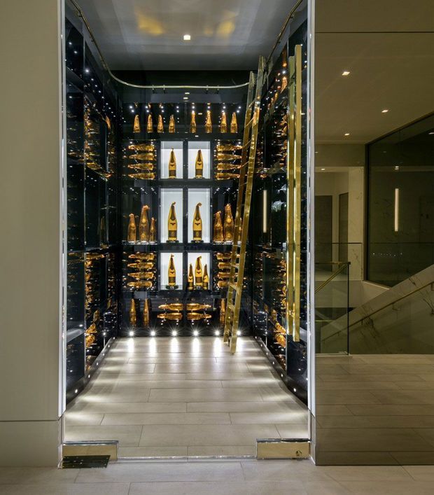A luxurious illuminated wine and champagne display room with glossy black mirrored walls, neatly arranged bottles on illuminated shelves, and a gold-toned leaning ladder. The compact room has large-format light grey floor tiles, recessed and shelf lighting, and opens onto a refined contemporary hallway with a glass balustrade and staircase.