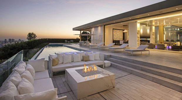 A luxurious contemporary rooftop terrace at dusk, featuring a large outdoor lounge with pale upholstered seating, a square built-in fire table, stepped decking, a glass balustrade, and a long cantilevered pavilion. An infinity-edge pool, sun loungers, and an open-plan illuminated interior are visible beyond, with a distant urban skyline in the background.