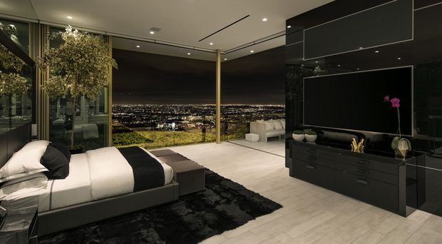 Luxurious contemporary bedroom with a low-profile upholstered bed, monochromatic black-and-white bedding, dark built-in media cabinetry, and a large television. Floor-to-ceiling glazing opens to expansive nighttime city views, while integrated ceiling lighting, a sculptural indoor tree, and polished dark finishes create a refined, dramatic atmosphere.