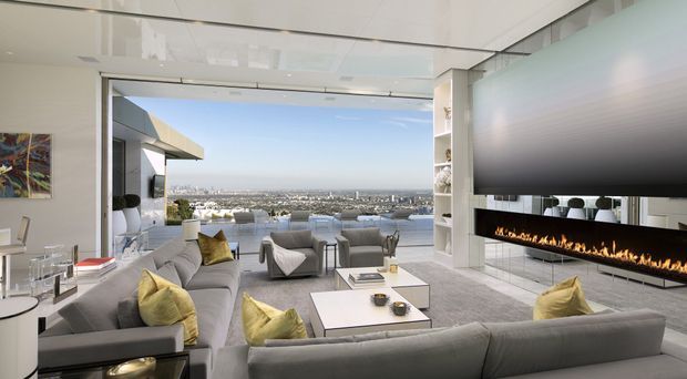 A luxurious contemporary living room with extensive floor-to-ceiling glazing opening onto a waterfront outlook. The room features large grey sectional sofas, pale yellow accent cushions, low white coffee tables, a pale grey rug, a sculptural lounge chair, built-in shelving, and a long linear fireplace beneath a large dark feature wall or television panel. The interior is bright and refined, with a wide view across water and a dense cityscape beyond.