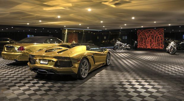 A dark, luxurious automotive showroom or private car gallery displaying several high-end sports cars and motorcycles. The space features a dramatic black-and-grey checkerboard floor, a low reflective ceiling with numerous recessed spotlights, dark walls and glazing, and a red geometric feature screen at the rear.