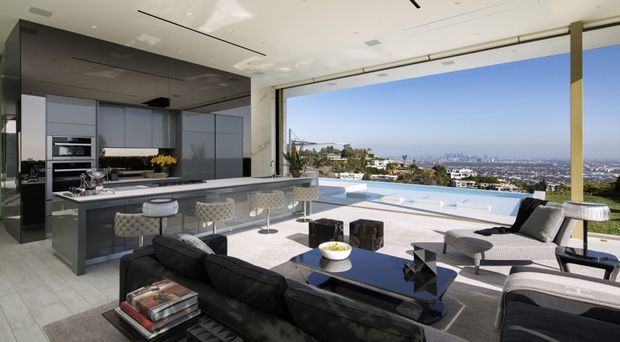 A luxurious open-plan indoor-outdoor living space with a sleek contemporary kitchen and island overlooking a panoramic city and coastal landscape. The room features floor-to-ceiling retractable glazing, a dark sectional sofa, sculptural lounge chairs, a reflective black coffee table, pale stone-look flooring, and a bright infinity pool terrace beyond.