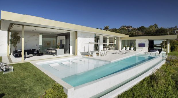 A low-slung modernist residence with extensive floor-to-ceiling glazing and deep covered outdoor living areas arranged around a long rectangular swimming pool. White rendered walls, flat roof planes, pale paving and minimalist detailing create a bright, geometric composition, with landscaped grounds and trees surrounding the property.