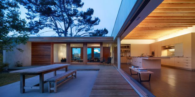 A sophisticated modern courtyard connecting an open-plan kitchen and living area with a timber-lined covered terrace. Floor-to-ceiling glazing opens toward a landscaped courtyard, while a long timber dining table, built-in kitchen cabinetry, warm concealed lighting, and exposed timber ceiling battens create a calm indoor-outdoor living space.