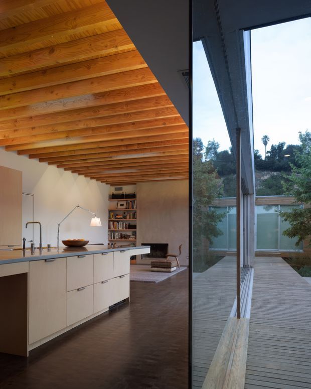 Interior view of a warm, minimalist kitchen opening into a living area, with a large central island, pale built-in cabinetry, dark timber flooring, and a dramatic exposed timber slatted ceiling. Full-height glazing beside the kitchen looks onto a timber deck and planted courtyard.