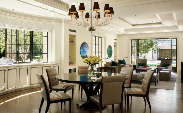 Bright, refined open-plan dining and living area with a large round dark dining table surrounded by upholstered chairs, adjacent built-in cabinetry and display shelving, and a lounge seating arrangement beyond. The room features large black-framed windows, layered coffered and tray ceiling detailing, a statement multi-arm chandelier, pale walls, and warm timber flooring.