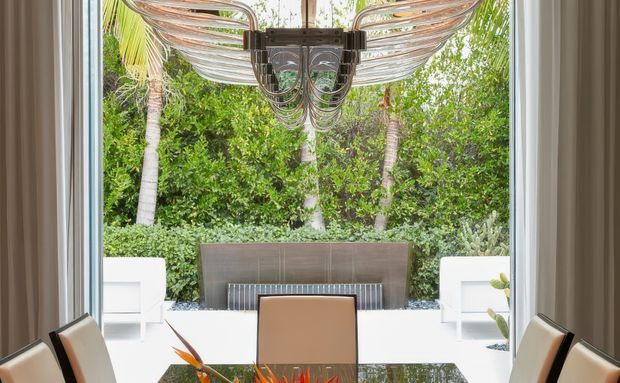 A bright, refined dining area viewed through large floor-to-ceiling glazing, with a reflective dark dining table, tan upholstered dining chairs, and an oversized sculptural metallic chandelier. The glazing opens visually onto a lush tropical garden with palms, white outdoor seating, and a linear water or fire feature beyond.