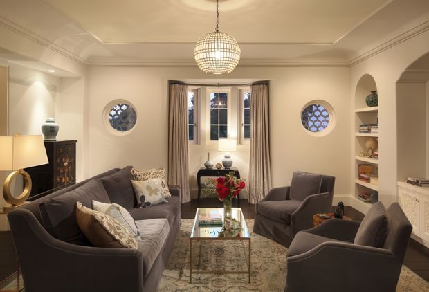A refined, traditionally styled living room with cream walls, decorative ceiling mouldings, a central crystal pendant light, and symmetrical circular windows with patterned glazing. Multiple deep grey upholstered sofas and armchairs surround a glass coffee table over a patterned area rug. Built-in arched shelving and decorative cabinetry frame the room, while tall curtains surround a central multi-pane window seat.