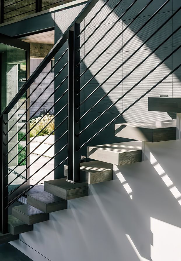 A bright, modern interior stairway with floating timber treads cantilevered from a white structural wall. The staircase features a prominent black metal central support and diagonal cable railing, with large glazed openings and strong sunlight casting geometric shadows across the walls and stairs. A landscaped exterior courtyard is visible beyond the glazing.