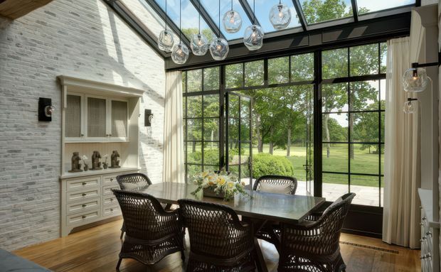 A bright dining room with a long dark dining table and woven chairs, positioned beside expansive black-framed floor-to-ceiling glazing. The room features a dramatic glazed roof or skylight structure, pale painted brick walls, timber flooring, built-in white cabinetry, and clustered glass pendant lights. Views through the glazing show a landscaped garden and lawn.