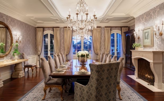 A formal, richly decorated dining room with a long dark wood dining table, upholstered dining chairs, patterned area rug, ornate chandelier, layered curtains, patterned wallpaper, decorative wall sconces, and a classic fireplace. The room has polished dark timber flooring, elaborate coffered ceiling detailing, and tall glazed doors/windows opening to a blue-toned exterior view.
