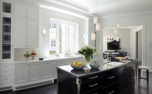 Bright, sophisticated kitchen with extensive pale grey built-in cabinetry, a large black island with drawers and a dark stone benchtop, pendant lights, a window over the sink, and integrated appliances. The room has classic panelled joinery and trim paired with contemporary fittings and high-contrast finishes.