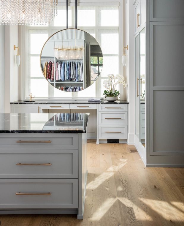 Bright, refined walk-in wardrobe with extensive pale grey built-in cabinetry, brass linear handles, dark stone benchtops, light timber flooring, and a large round mirror reflecting hanging clothes and a decorative chandelier. Tall windows provide abundant natural light, while a central island offers additional drawer storage and surface space. The room has a polished, contemporary-classic appearance with elegant detailing.