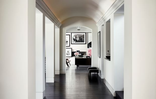 A long, bright interior hallway with a curved barrel-vaulted ceiling, white walls and built-in openings, dark timber flooring, and black-and-white furnishings. The corridor leads toward a styled sitting or study area with framed artwork and a dark console or desk.