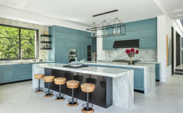 A spacious, refined kitchen with blue built-in cabinetry, a large white-and-grey veined stone island and matching splashback, black accents, and five bar stools. A linear geometric pendant hangs above the island, while large windows bring in greenery and natural light. Decorative wall panelling and marble-look surfaces create a polished contemporary-classic feel.