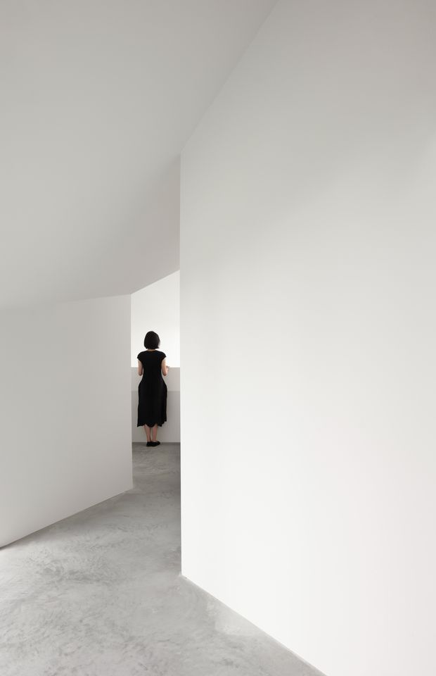 A minimalist white interior corridor or circulation space with angular, sculptural walls and ceiling planes. The floor is a smooth mottled grey concrete-like finish, while a person in dark clothing stands in the distant opening. The composition is sparse, geometric, and strongly monochromatic with soft natural light.